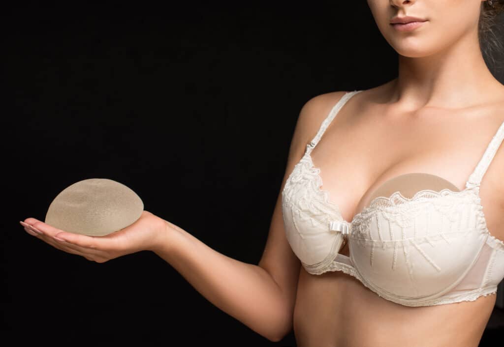 Woman in a white bra holding a silicone breast implant in her hand, with a second implant placed inside the bra