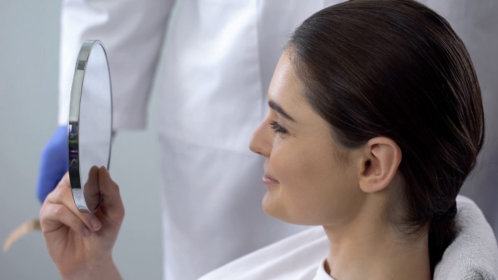 Woman looking in the mirror feeling happy seeing final results of her revision rhinoplasty